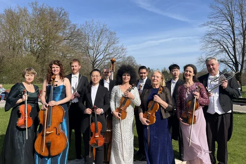 Musikgruppe im Park Eine Gruppe von zwölf Musikern steht im Freien, umgeben von Bäumen. Sie halten Instrumente wie Geigen, Cello und Trompete. Die Musiker sind in formeller Kleidung und lächeln in die Kamera.