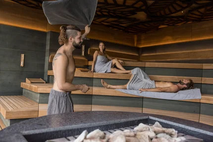 Entspannung in der Sauna Eine sauna mit drei Personen: Ein Mann schwenkt ein Handtuch, während zwei andere entspannt auf Bänken liegen. Es gibt einen Steingrill in der Mitte.