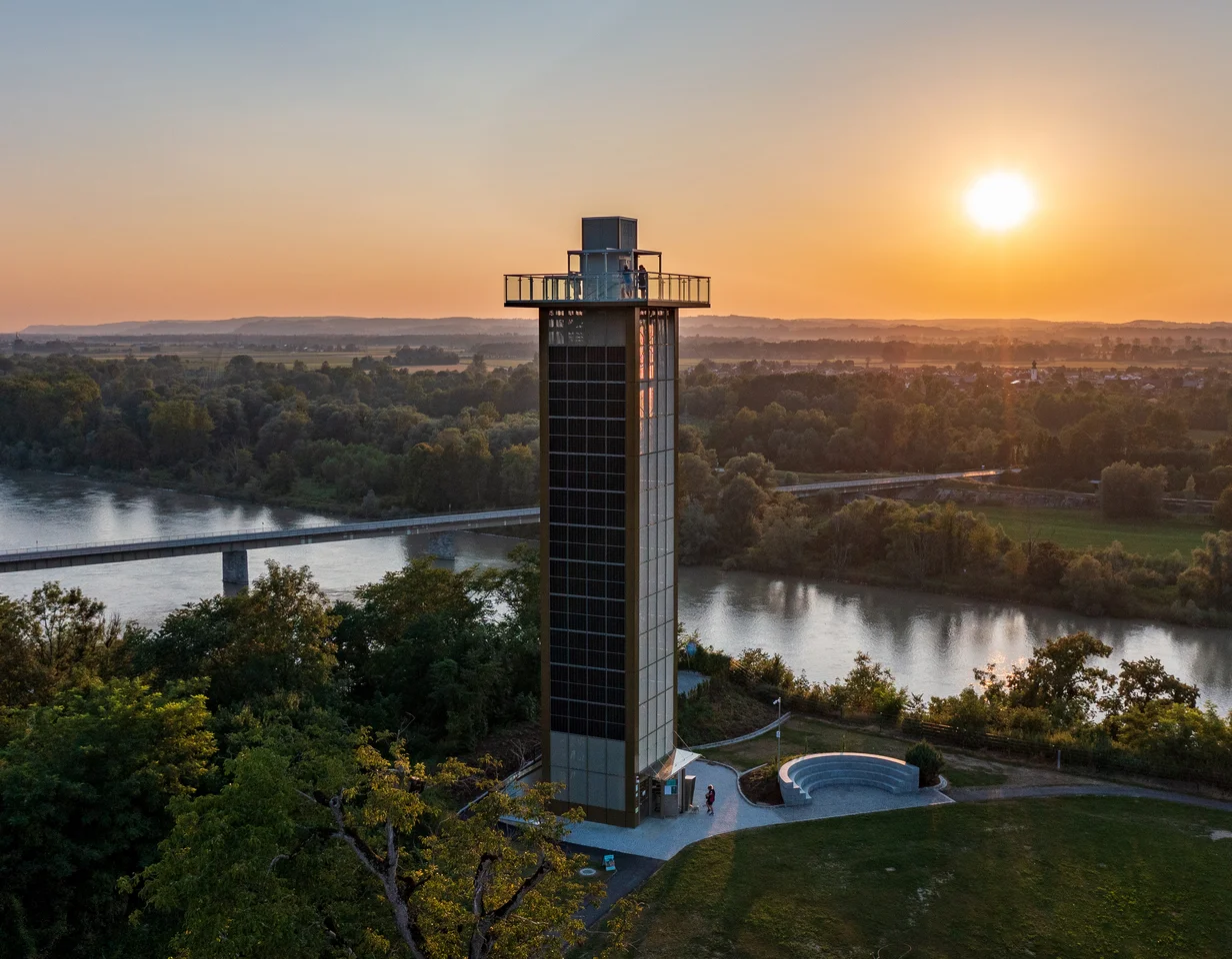 Ein Aussichtsturm steht am Ufer eines Flusses bei Sonnenuntergang. Umgeben von Bäumen und grünen Wiesen, mit einer Brücke im Hintergrund.