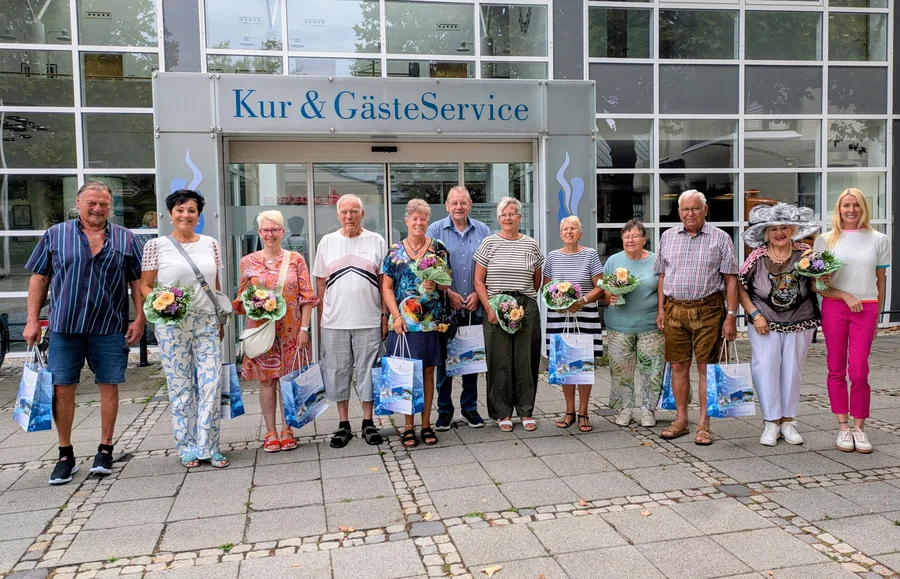 Eine Gruppe von zehn Personen steht vor einem Empfangsgebäude mit dem Schriftzug 'Kur & GästeService'. Jeder hält eine Blume oder einen Strauß, neben einigen Taschen.