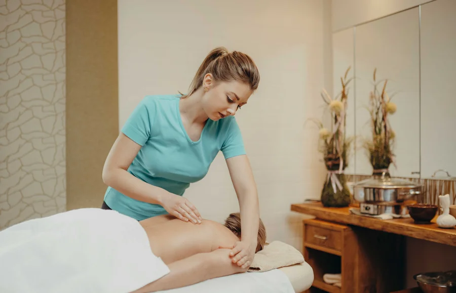 Massage in einem Spa Eine Masseurin führt eine Massage durch. Die Klientin liegt auf einer Liege, mit einem Handtuch bedeckt, während die Masseurin ihren Rücken massiert. Im Hintergrund sieht man Pflanzen und Massageutensilien.