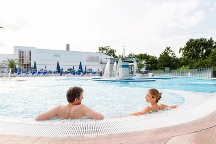 Ein Mann und eine Frau entspannen im Wasser eines Pools, umgeben von Sonnenliegen und Bäumen. Es gibt einen Wasserfall im Hintergrund und hellen Himmel.