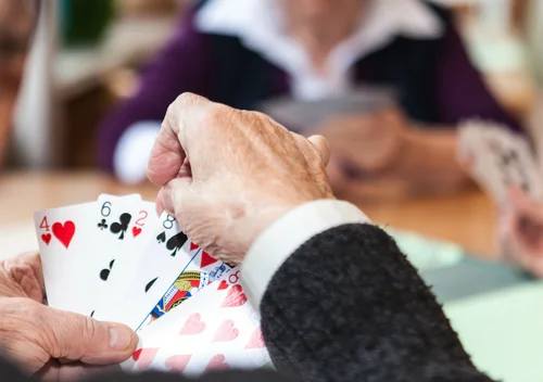 Spielen von Karten Eine Hand hält Spielkarten mit verschiedenen Farben und Zahlen, während im Hintergrund andere Spieler zu sehen sind, die ebenfalls Karten halten.