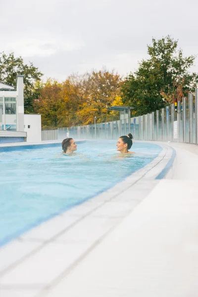 Two women relax in the warm water of a pool, surrounded by autumn trees in the background. There is a calm, relaxing atmosphere with light steam over the water.