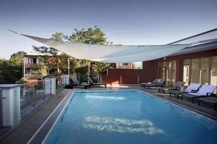 Ein Schwimmingpool mit Liegestühlen unter einem Sonnensegel, umgeben von Bäumen und einem Gebäude im Hintergrund.