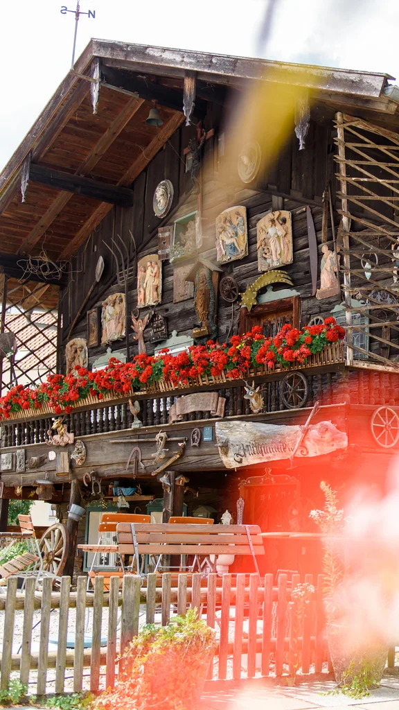 Ein traditionelles Holzhaus mit blühenden Fensterkästen und verschiedenen dekorativen Elementen. Tische und Stühle auf dem Grundstück sind sichtbar, umgeben von einem Holzzaun.