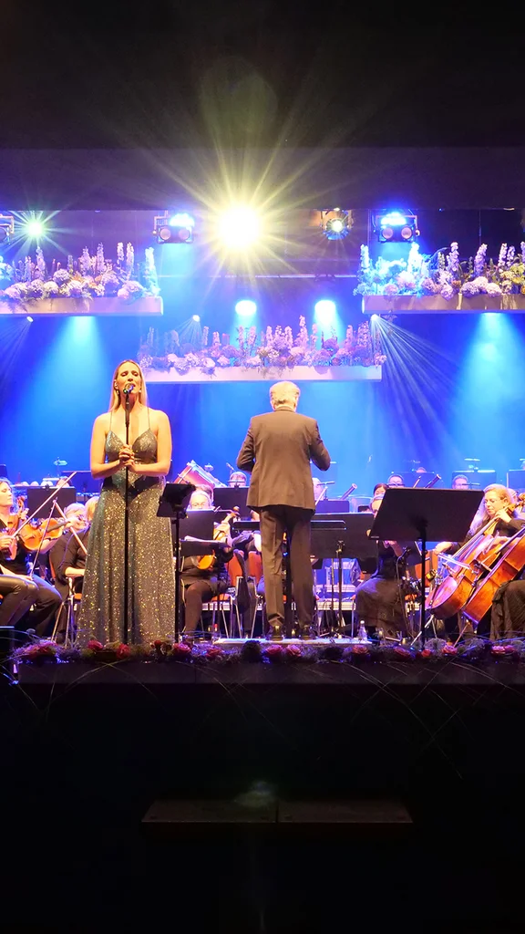 A live music performance featuring female vocalists, an orchestra, and musicians on stage. Bright lights illuminate the scene, creating a vibrant atmosphere. The audience is visible in the foreground, enjoying the concert.