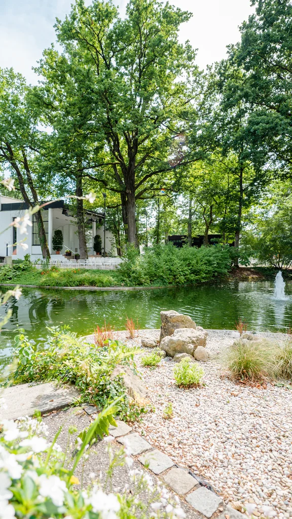 Ruhiger Teich mit Springbrunnen Ein ruhiger Teich umgeben von Bäumen und Blumen, mit einem kleinen Springbrunnen in der Mitte. Im Hintergrund ist ein modernes Gebäude sichtbar.