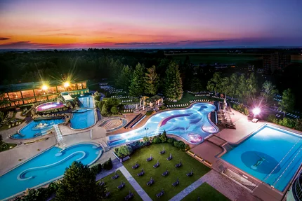 Ein Panoramablick auf ein Freibad mit mehreren Pools, umgeben von Bäumen und Liegestühlen, unter einem bunten Sonnenuntergang.