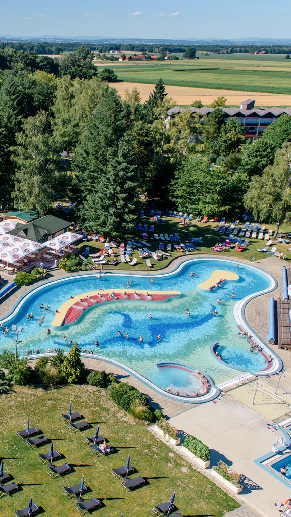 Wasserpark mit Schwimmbädern Ein großer Wasserpark mit mehreren Becken, Rutschen und Liegen. Menschen schwimmen in verschiedenen Bereichen, während im Hintergrund Bäume und Wiesen sichtbar sind.