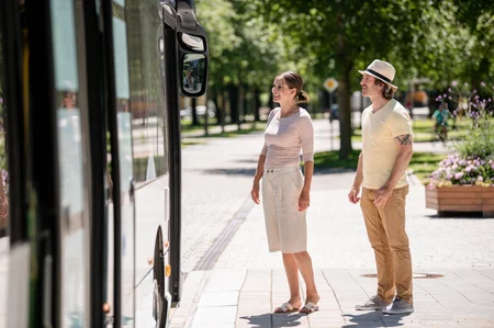 Warten auf den Bus Zwei Personen stehen an einer Bushaltestelle und schauen auf den Bus. Eine Frau lächelt, ein Mann beobachtet sie.