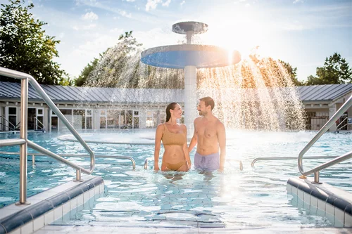 Paar im Schwimmbecken Ein Paar steht im Wasser eines Schwimmbeckens, umgeben von sprudelndem Wasser und Bäumen im Hintergrund. Ihre Gesichter zeigen Freude und Zuneigung zueinander.