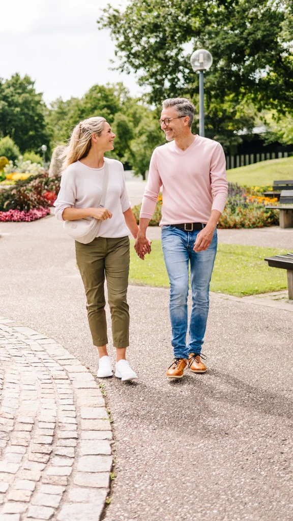 Couple Walking in Park A smiling middle-aged couple walks hand in hand along a paved path in a park beside a fountain and colorful flower beds, with benches and trees around them.
