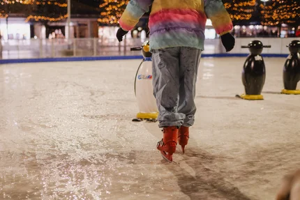 Person beim Eislaufen Eine Person in bunter Winterjacke läuft auf einer Eisbahn mit roten Schlittschuhen; im Hintergrund stehen Pinguin-Lernhilfen und eine Eishallenbegrenzung.