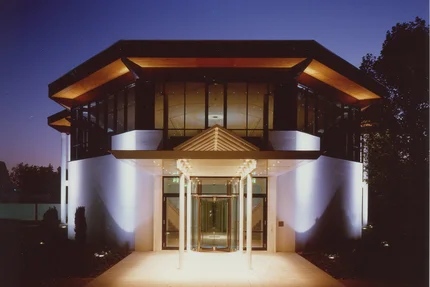 Modernes Gebäude bei Nacht Ein modernes Gebäude mit gläserner Fassade, beleuchtet bei Nacht, mit einem Zeltdach über dem Haupteingang und Plattform aus Fliesen.