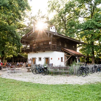 Ein rustikales Gebäude, umgeben von Bäumen, mit einer Sonneneinstrahlung. Im Vordergrund stehen Fahrräder, und Menschen essen und trinken an einer Freiluftterrasse.