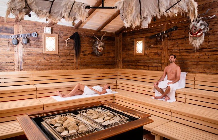 Ein Saunaraum mit Holzwänden und Holzbänken. Zwei Personen, eine liegend und eine sitzend, tragen Handtücher. Über ihnen hängen dekorative Tierköpfe und ein Herd mit Steinen ist in der Mitte.