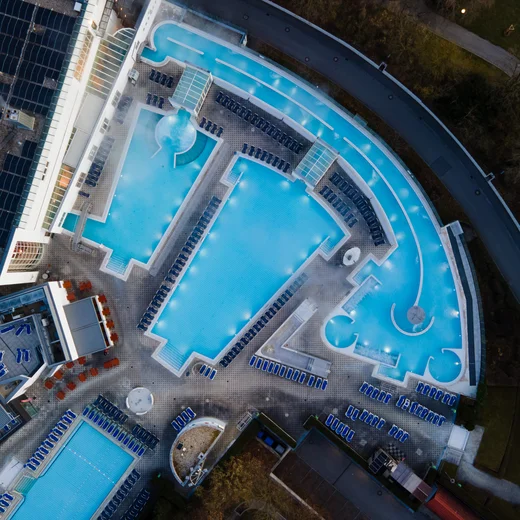 Luftaufnahme eines Schwimmbadkomplexes Vogelperspektive auf ein großes Schwimmbadgelände mit mehreren blauen Pools und Liegen. Die Umgebung enthält auch einige Gebäude und eine grüne Rasenfläche.