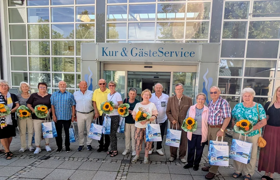 Eine Gruppe älterer Menschen und eine jüngere Frau steht vor dem Eingang von „Kur & GästeService“, mehrere halten Sonnenblumensträuße und Einkaufstaschen.