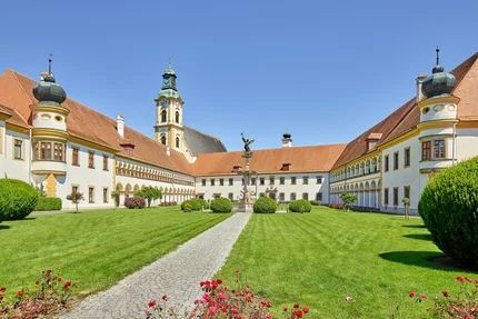 Ein weitläufiger Innenhof eines historischen Gebäudes mit grünem Rasen, Rosenbeeten und einer Statue in der Mitte. Umgeben von klassischer Architektur mit Türmchen.