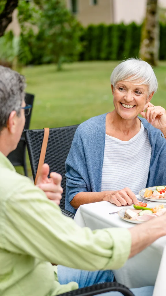 Ein älteres Paar sitzt an einem Tisch im Freien mit Essen und Getränken. Die Frau lächelt und hört dem Mann zu, der eine handgestellte Geste macht. Um sie herum ist eine grüne Umgebung und ein schöner Garten.