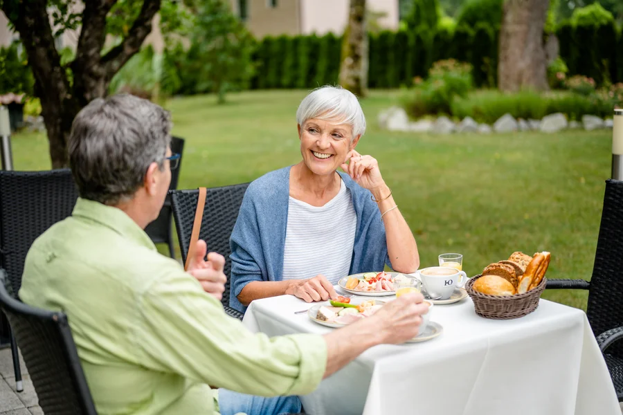 Ein älteres Paar sitzt an einem Tisch im Freien mit Essen und Getränken. Die Frau lächelt und hört dem Mann zu, der eine handgestellte Geste macht. Um sie herum ist eine grüne Umgebung und ein schöner Garten.
