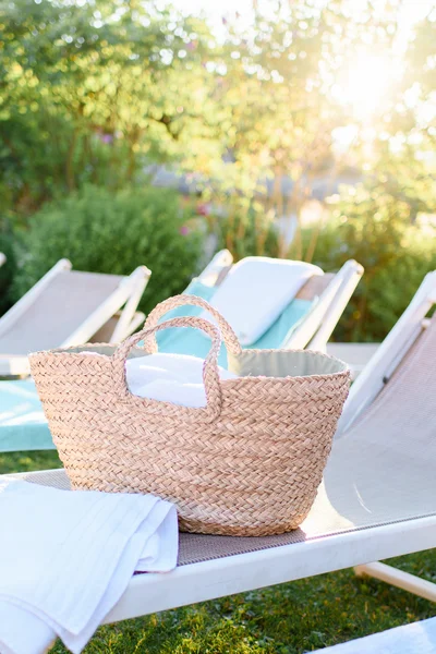 A woven basket with towels rests on a lounge chair in a garden with sunlight. In the background, more lounge chairs and colorful flowers are visible.