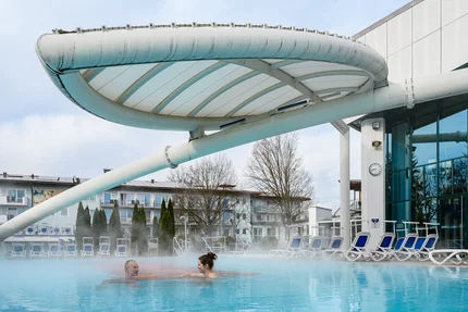 Außenpool mit Rutsche Ein Mann und eine Frau schwimmen in einem Außenpool mit aufsteigendem Dampf; daneben stehen Liegen, und darüber ragt eine große überdachte Rutsche oder Konstruktion aus dem Gebäude.