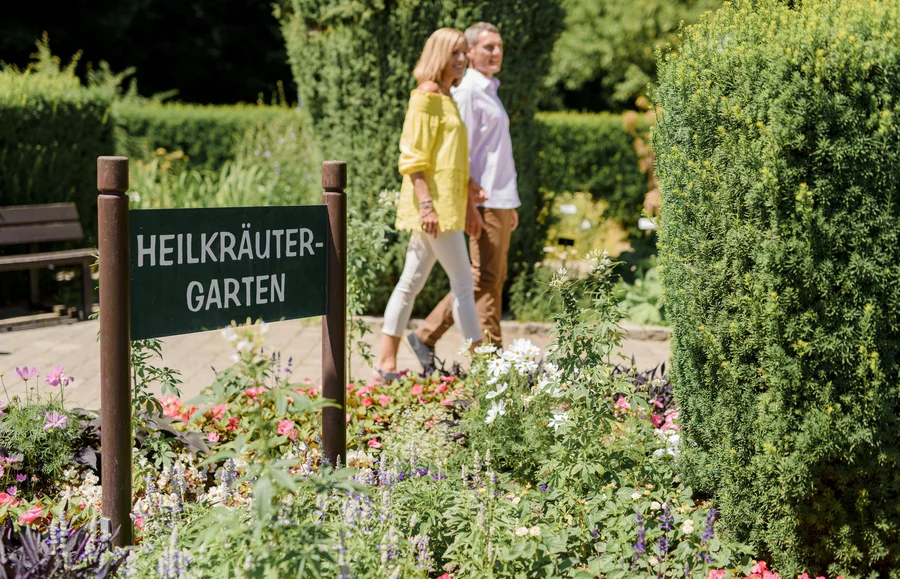 Zwei Personen gehen in einem Kräutergarten, während ein Holzschild mit der Aufschrift 'Heilkrautgarten' im Vordergrund steht. Bunte Blumen umgeben den Weg.