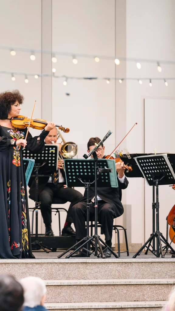 Eine Musikerin spielt Violine auf einer Bühne, umgeben von einem Ensemble. Im Hintergrund sind mehrere Musiker mit Notenständern zu sehen. Der Veranstaltungsort ist ein geschmückter Bereich mit Pflanzen und Lichtern.