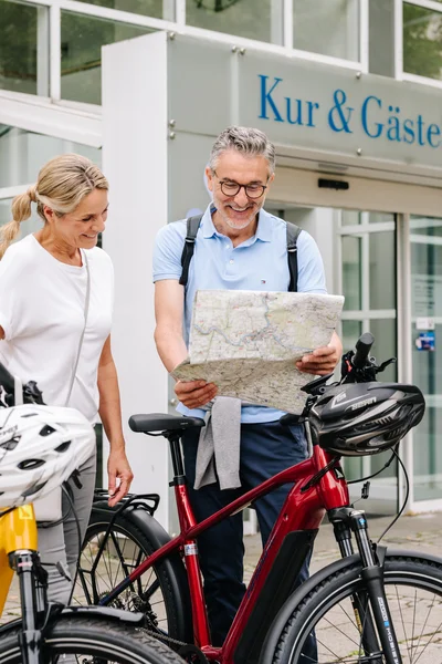 Ein Mann und eine Frau stehen mit zwei Fahrrädern vor einem Gebäude. Der Mann hält eine Karte, während die Frau lächelt. Die Szene zeigt regionale Gastfreundschaft.