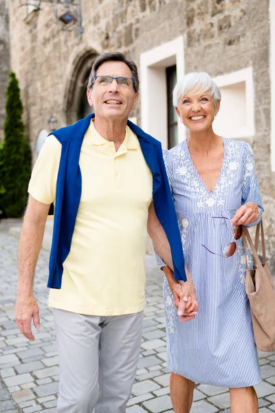 Ein älteres Paar läuft Hand in Hand auf einem gepflasterten Weg, umgeben von grünen Büschen und alten Gebäuden. Beide lächeln, der Mann trägt ein gelbes Shirt mit einer blauen Weste und die Frau ein gestreiftes Kleid.