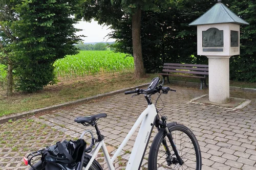 Ein E-Bike steht auf einem Pflasterweg, umgeben von Bäumen und einer Bank. Im Hintergrund ist ein Feld zu sehen.