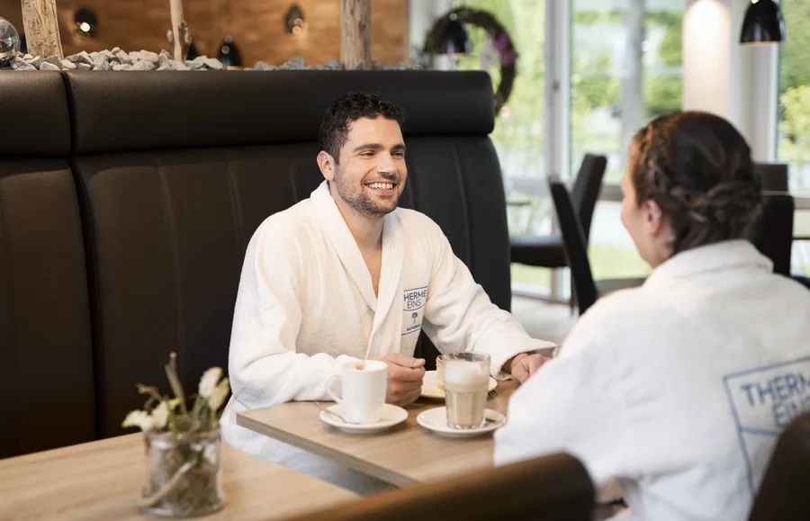 Ein Mann und eine Frau in weißen Bademänteln sitzen an einem Tisch, genießen Getränke und lächeln einander zu. Auf dem Tisch stehen zwei Tassen Kaffee.