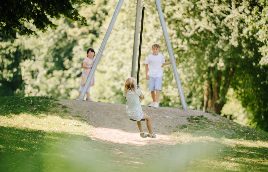 Ein kleines Kind auf einer Schaukel, während zwei andere Kinder im Hintergrund stehen und zuschauen. Die Szene spielt sich in einem parkähnlichen Umfeld mit Bäumen ab.