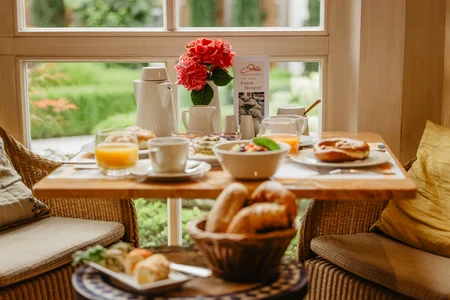 Frühstückstisch mit verschiedenen Speisen Ein Tisch mit einem Frühstück, das frische Brötchen, Salat, Obstschüssel, Joghurt und Getränke wie Kaffee und Saft beinhaltet. Im Hintergrund sind grüne Pflanzen sichtbar.