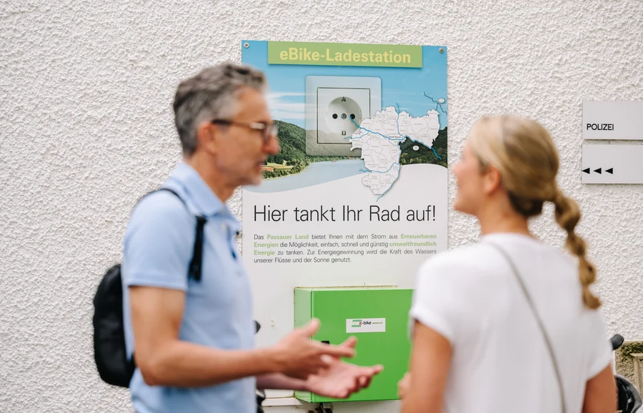 Ein Mann und eine Frau stehen vor einer eBike-Ladestation an einer Wand. Auf dem Schild steht „Hier tankt Ihr Rad auf!“; daneben hängt ein Schild mit „Polizei“.