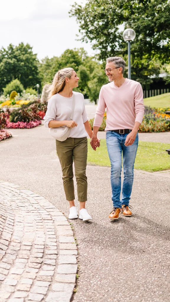 Paar im Park spazieren Ein Paar geht Hand in Hand entlang eines gepflasterten Weges in einem Park. Blumenbeete und Bänke sind im Hintergrund zu sehen.