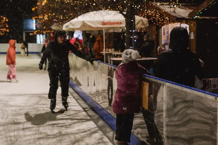 Eisbahn bei Nacht Kinder und Erwachsene laufen auf einer beleuchteten Eisbahn im Schnee, neben der Absperrung stehen Personen, im Hintergrund sind ein geschmückter Baum und ein Verkaufsstand zu sehen.