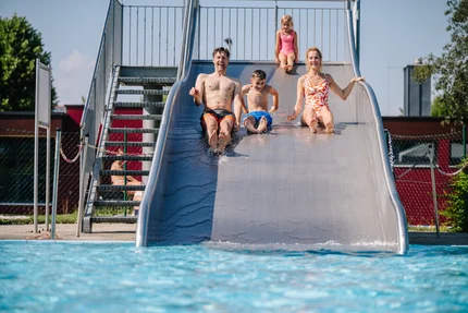 Eine Familie rutscht gemeinsam von einer Rutsche ins Wasser des Schwimmbads. Es sind ein Vater, zwei Kinder und eine Mutter zu sehen. Sie wirken fröhlich und es spritzt Wasser.