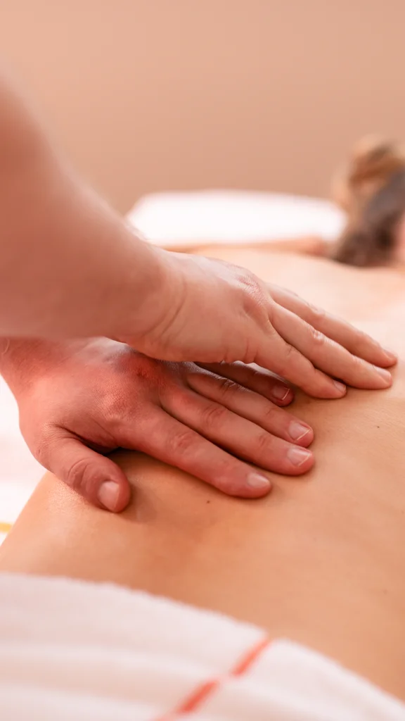 Eine Person erhält eine Rückenmassage. Ein Masseur drückt seine Hände sanft auf den Rücken der liegenden Person, die mit einem Handtuch bedeckt ist. Der Raum wirkt entspannend und hell.
