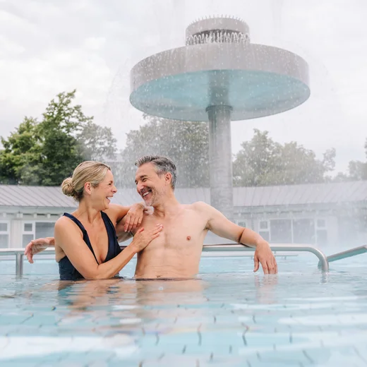 Ein Paar sitzt in einem Schwimmbecken und lächelt sich an. Im Hintergrund ist ein großer Wasserfall zu sehen. Bäume umgeben den Bereich.