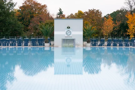 Blick über ein Außenbecken der Europa Therme mit Liegestühlen, Palmen und herbstlich gefärbten Bäumen im Hintergrund.