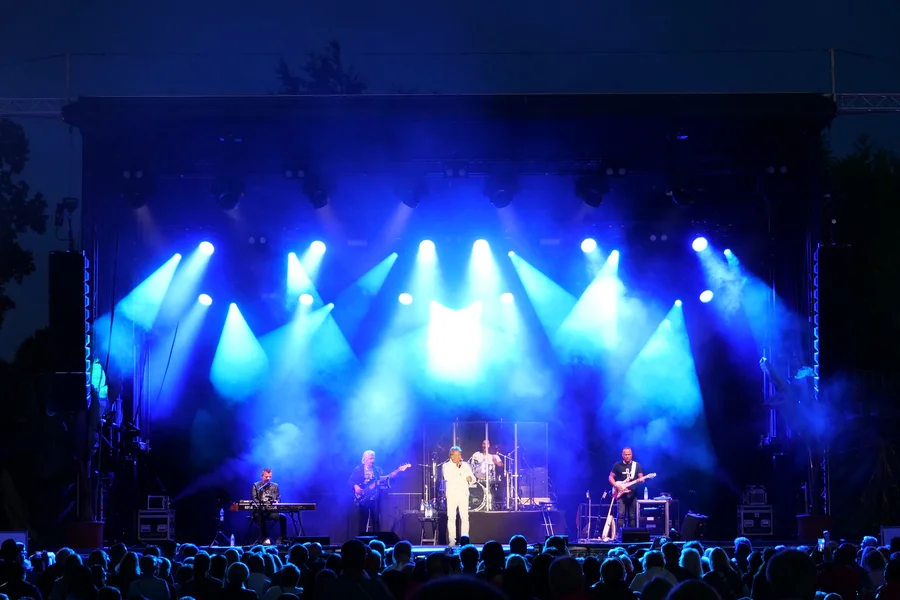 Ein Sänger in weißem Anzug tritt auf einer großen Open-Air-Bühne auf, flankiert von zwei Leinwänden. Blaue Scheinwerfer beleuchten die Band, und vor der Bühne steht ein dichtes Publikum.