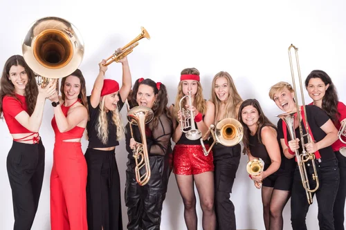 Musikerinnen mit Blasinstrumenten Eine Gruppe von Musikerinnen in bunten Outfits hält verschiedene Blasinstrumente, darunter Posaunen und Trompeten. Sie stehen fröhlich in einer Reihe und lächeln. Die Farbpalette umfasst Rot, Schwarz und einige glitzernde Elemente.
