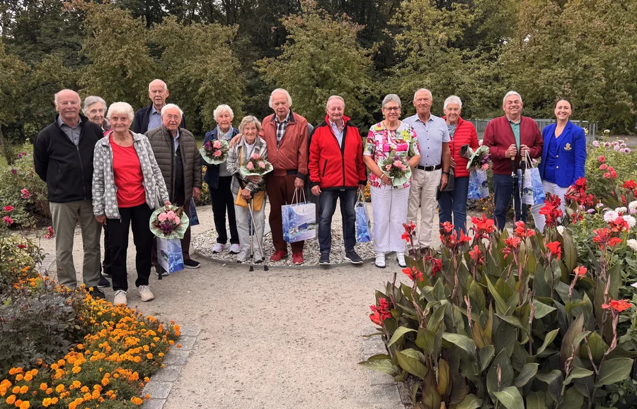 Eine Gruppe älterer Menschen steht auf einem Gartenweg zwischen Blumenbeeten und hält Sträuße sowie Geschenktüten in den Händen.