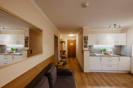 Ein Innenraum zeigt eine gepolsterte Couch, eine Küchenzeile mit weißen Möbeln, einem Tisch, Stühlen und einem Eingang. An der Wand hängt ein Spiegel, und im Hintergrund sind einige Türen zu sehen.