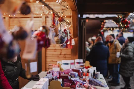 Holiday Market Stall A festive market stall with various holiday ornaments and gift items displayed. People are browsing in the background, surrounded by twinkling lights.