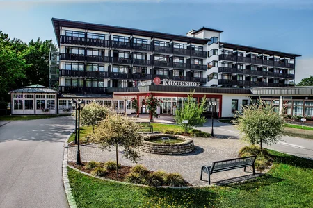 Hotel Königshof Außenansicht Dies ist ein Hotel namens Königshof, das von einem gepflegten Garten umgeben ist. Im Vordergrund befindet sich ein gepflasterter Bereich mit einer kleinen Fontäne und Bänken. Das Gebäude hat Balkone und große Fenster.