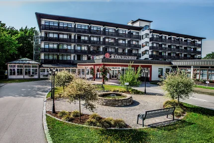 Hotel Königshof Außenansicht Dies ist ein Hotel namens Königshof, das von einem gepflegten Garten umgeben ist. Im Vordergrund befindet sich ein gepflasterter Bereich mit einer kleinen Fontäne und Bänken. Das Gebäude hat Balkone und große Fenster.